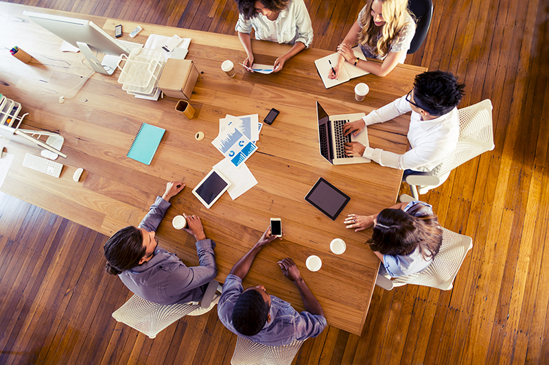 Aerial view of designers and engineers during a design meeting. The large wooden table around which they are seated contains sketches and notes as well as a series of laptops and mobile devices, such as tablets and smartphones.